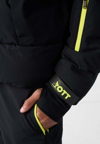 Zwarte donsjas met felgele ritsen en accenten. Voorzien van een logo op de boord en een ritszak met een bijpassende rand.