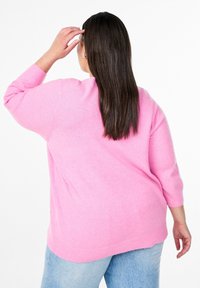 Hellrosa Pullover mit dreiviertel Ärmeln und geripptem Saum. Weiche Textur, lockere Passform. Getragen über hellblauen Jeans.