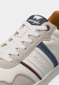 Λευκό αθλητικό παπούτσι Wrangler με μπεζ σουέντ λεπτομέρειες, μπλε ναυτικό ρίγα, λευκά κορδόνια και καφέ εσωτερική επένδυση, που δείχνει τα λογότυπα της μάρκας στη γλώσσα και στην πλευρά.