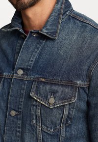 Osoba s plnovousem nosící modrou džínovou bundu s kovovými knoflíky a náprsní kapsou, na které je štítek "Polo".