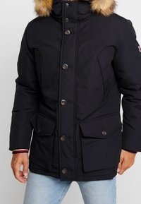 Sort parka-jakke med hætte foret med kunstpels, trykknapper og knapper, to forlommer og en skræddersyet pasform. Har et letvægtsdesign.