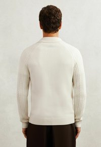 Weißer Pullover mit einem strukturierten Zopfmuster an den Ärmeln, gerippten Ärmelbündchen und Saum sowie einer taillierten Passform. Solide Rückansicht.