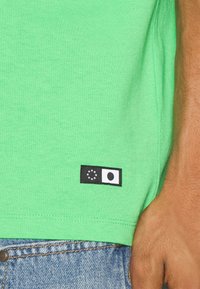 Zielony bawełniany t-shirt z okrągłym dekoltem, z czarną metką zawierającą dwa logo na dolnym brzegu. Tkanina jest gładka i lekka.
