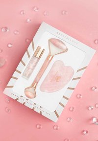 Zestaw do pielęgnacji twarzy z kamieniami szlachetnymi w pudełku z różowymi złotymi akcentami. Zawiera wałek, narzędzie gua sha i serum w przezroczystym pojemniku. Różowe tło.