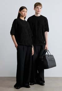 Μαύρα πλεκτά πουλόβερ με υφή και κοντά μανίκια, συνδυασμένα με φαρδιά μαύρα παντελόνια. Μια μαύρη τσάντα κρατείται από μια φιγούρα, τονίζοντας τον μινιμαλιστικό σχεδιασμό.