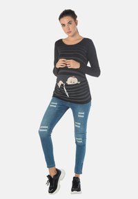 Schwarzes Langarmoberteil mit horizontalen Streifen und einem cartoonhaften Babydesign auf der Vorderseite, kombiniert mit ausgebleichten blauen Jeans und schwarzen Turnschuhen.