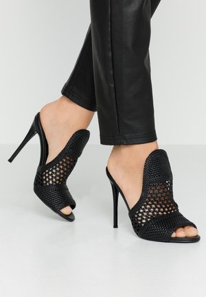 Feet in black woven mesh high-heeled mules with open toes, paired with black fitted pants against a plain light background.