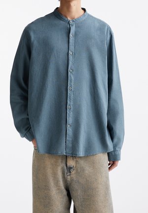 Homme portant une chemise bleue délavée ample à boutons avec col mandarin et pantalon beige délavé, mains dans les poches, fond uni.