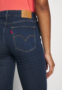 Persona que lleva vaqueros Levi's azul oscuro con bolsillo trasero cosido y parche de cuero de la marca, combinados con una camisa coral.