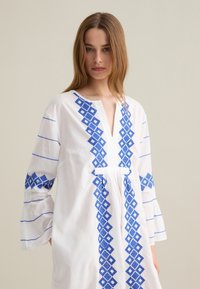 Tunic bianca con ricami a diamante blu lungo il davanti e le maniche. Presenta un collo a V, maniche lunghe e polsini svasati.