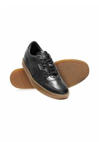 Czarne skórzane sneakersy z gumową podeszwą, z sznurowadłami i detalami w formie przeszyć, pokazane z jednym butem stojącym, a drugim na boku z podeszwą do góry.