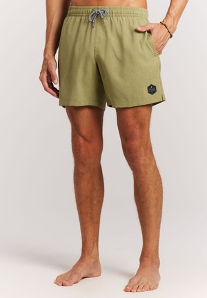 Man draagt olijfgroene zwembroek met zwart-wit trekkoord, staat blootsvoets met één hand in de zak tegen een effen achtergrond.