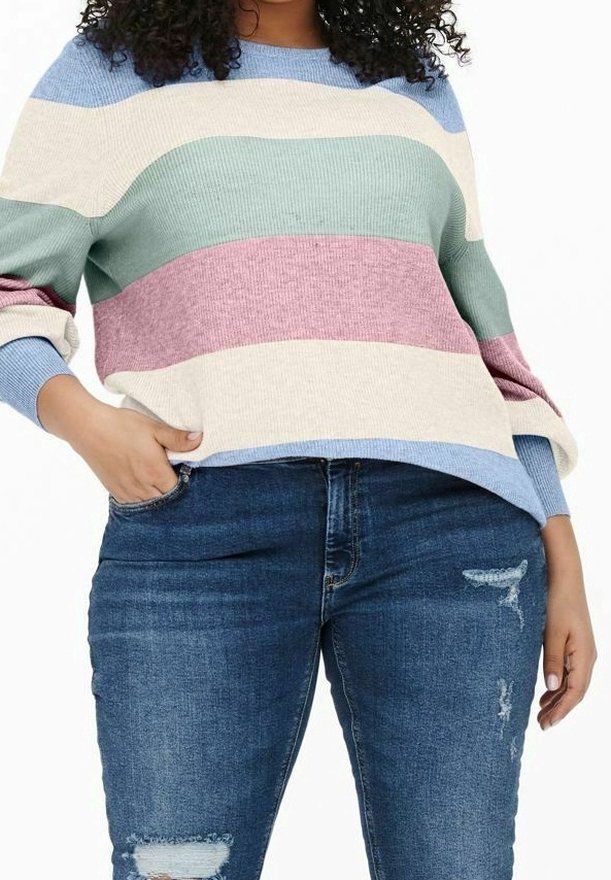 Personne aux cheveux bouclés portant un pull rayé aux tons pastel et un jean bleu délavé, une main dans la poche et l'autre détendue.