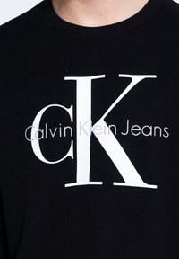 T-shirt nera in cotone con grande logo bianco "CK" e testo "Calvin Klein Jeans", design con collo rotondo, texture liscia e vestibilità casual.