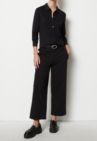 Svart långärmad skjorta med knappar, kombinerad med vida svarta byxor och kraftiga svarta loafers. Ett smalt bälte med ett runt spänne fulländar looken.