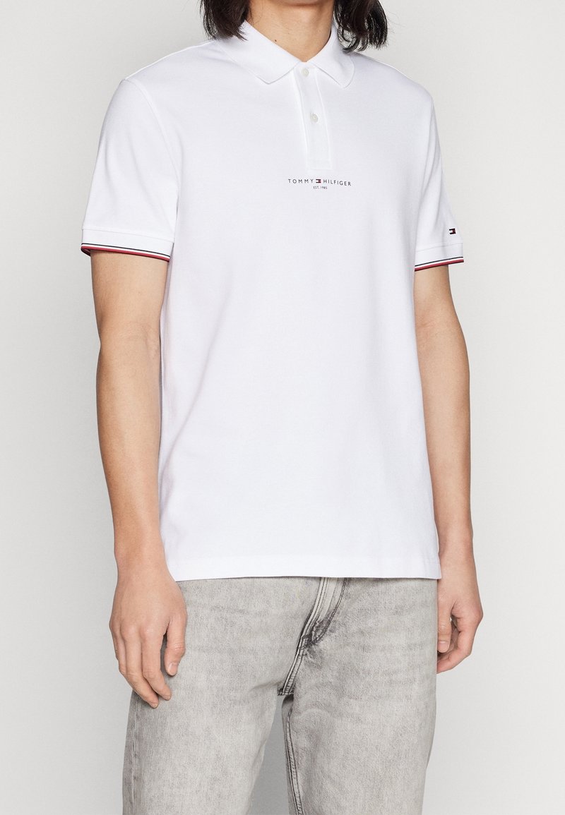 Homme portant un polo blanc Tommy Hilfiger avec le logo sur la poitrine et la manche, associé à un jean gris clair, debout devant un fond uni.