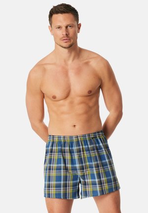 Mann mit kurzem Haar, der blaue, grüne und gelbe karierte Boxershorts trägt und mit hinter dem Rücken verschränkten Armen vor einem schlichten Hintergrund steht.