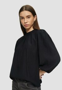 Schwarze, strukturierte Bluse mit höherem Kragen und voluminösen, puffigen Ärmeln. Verfügt über Falten an den Schultern. Getragen mit dunklen Jeans.