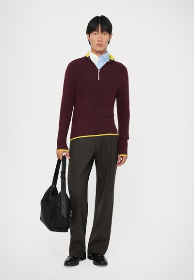 Jersey burdeos con detalles amarillos, cuello con cremallera, combinado con pantalones marrones de pierna ancha; la modelo lleva un bolso negro al hombro.
