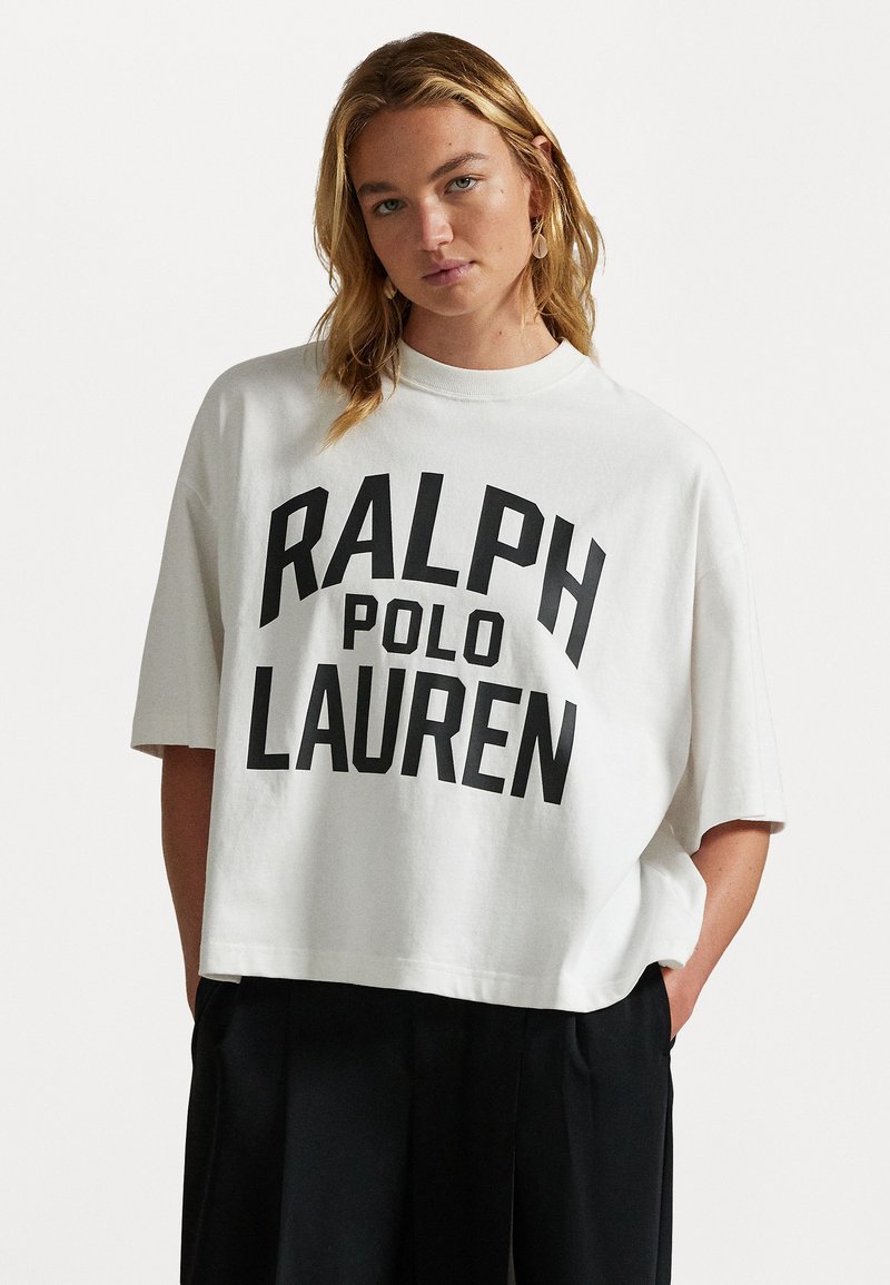 Balta kokvilnas T-kreklu ar brīvu piegriezumu, ar melnu pārāk lielu "RALPH POLO LAUREN" grafiku priekšā. Īsās piedurknes un apaļa kakla izgriezums.