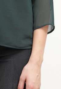 Gros plan du bras d'une personne portant un chemisier à manches transparentes vert foncé et un pantalon noir sur un fond clair.