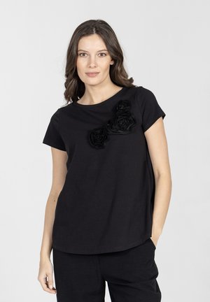 Kobieta o brązowych włosach do ramion, ubrana w czarną koszulkę z krótkim rękawem, ozdobioną trzema czarnymi dekoracjami w kształcie kwiatów na piersi.