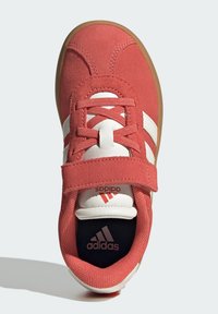 Červené semišové tenisky s bielymi pruhmi, suchým zipsom a čiernou vnútornou podrážkou s bielym logom. Okrúhly špička a gumová podrážka.