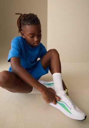 Fonott hajú gyerek kék ruhában a padlón ül, és fehér, zöld és kék csíkos sportcipőt kötöz fehér zokniban.