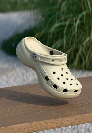 Beżowy klapek Crocs z otworami wentylacyjnymi i regulowanym paskiem unoszący się nad drewnianą powierzchnią, w tle zieleń.
