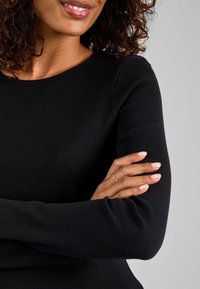 Vrouw met krullend haar die een zwarte top met lange mouwen draagt, licht glimlachend met gekruiste armen, en toont twee gouden ringen aan de vingers van haar linkerhand.