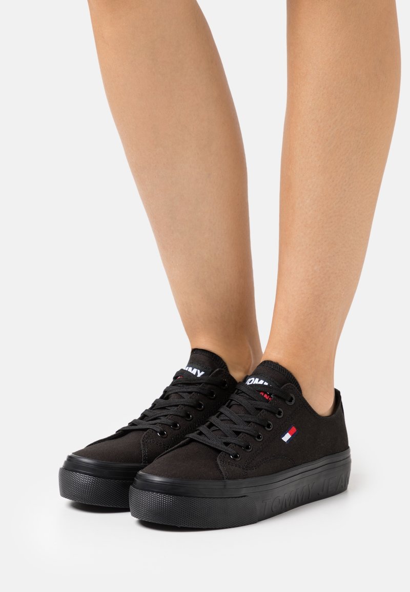 Personne portant des baskets basses noires avec des lacets noirs et un logo discret rouge, blanc et bleu sur le côté.