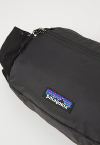 Schwarze Tasche mit glatter Textur, ausgestattet mit einem Reißverschluss und einem Patagonia-Logo-Patch mit mehrfarbigem Bergdesign.