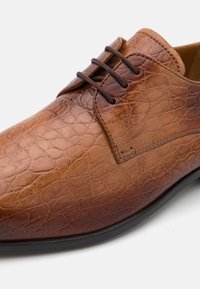 Zapato de cuero marrón con un acabado texturizado y agrietado, que presenta cordones redondos y sutiles detalles de costura, en una forma elegante con una suela negra.