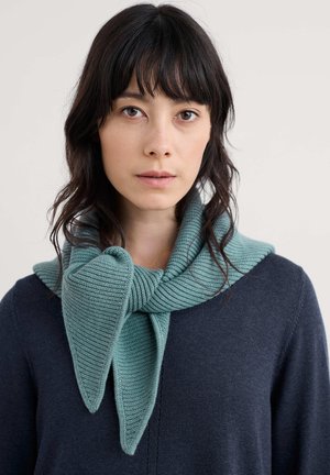 Strikket tørklæde i teal med et tekstureret, ribbet mønster. Har en stor sløjfedetalje og bæres over en mørk sweater. Glat og blødt udseende.