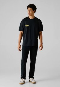 Noor mees seisab, kandes musta graafilise motiiviga t-särki, musti pükse ja valgeid tossusid, lihtsa helehall tausta ees.