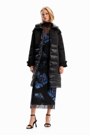 Doudoune noire avec fermeture éclair et capuche, portée sur une robe fleurie aux tons bleus. Le mannequin porte des sandales noires à talons.