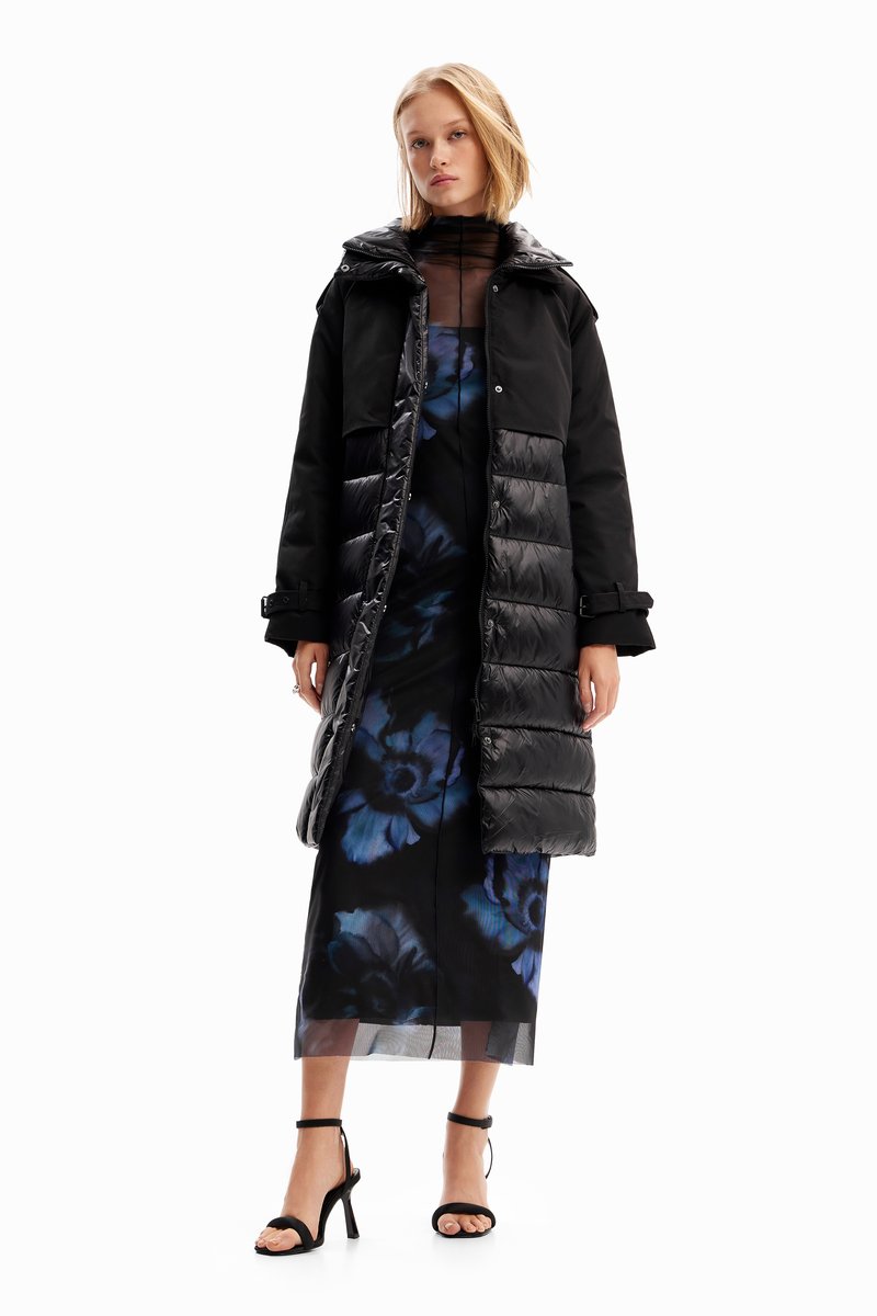 Doudoune noire avec fermeture éclair et capuche, portée sur une robe fleurie aux tons bleus. Le mannequin porte des sandales noires à talons.