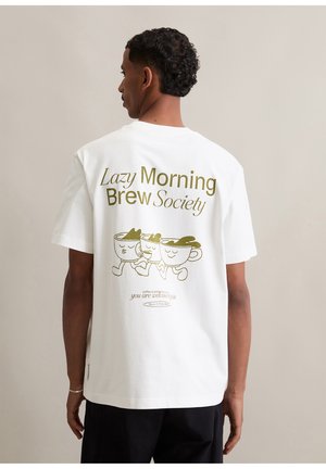 Man in een wit T-shirt met gouden tekst "Lazy Morning Brew Society" en een illustratie van drie lopende koffiekopjes op de achterkant.