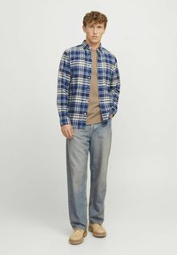 Blauw-wit geruite flanellen shirt over een beige t-shirt, gecombineerd met lichtblauwe jeans en tan instap schoenen.