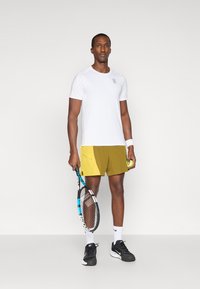 Άσπρη αθλητική μπλούζα, κίτρινες σορτς με πλαϊνές λεπτομέρειες, μαύρα αθλητικά παπούτσια, κρατώντας μια ρακέτα τένις και μια μπάλα. Απλός σχεδιασμός και κομψή εφαρμογή.