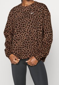 Bruine sweatshirt met lange mouwen en luipaardprint, met een wit Nike-logo op de borst. Zacht materiaal met een relaxte pasvorm. Zwarte leggings zichtbaar.
