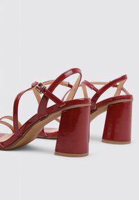Sandalias de tacón de charol rojo con punta abierta. Cuentan con correas entrecruzadas y un tacón grueso, destacando un acabado suave y brillante.