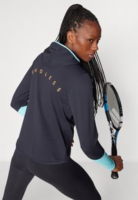 Tamno plava hoodie s svijetloplavim akcentima, s natpisom "ENDLESS" u narančastoj boji na leđima. Model drži tenisku reketa.