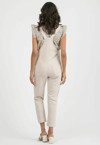 Beige cotton overalls with a fitted design, featuring pocket details and a floral print blouse with ruffled sleeves peeking out.