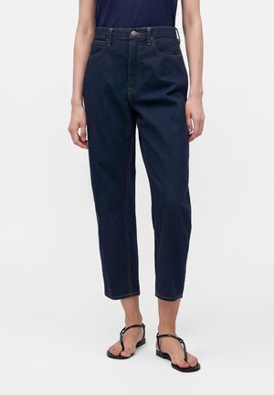 Personne portant un jean bleu foncé taille haute droit et des sandales plates noires à brides sur un fond blanc.