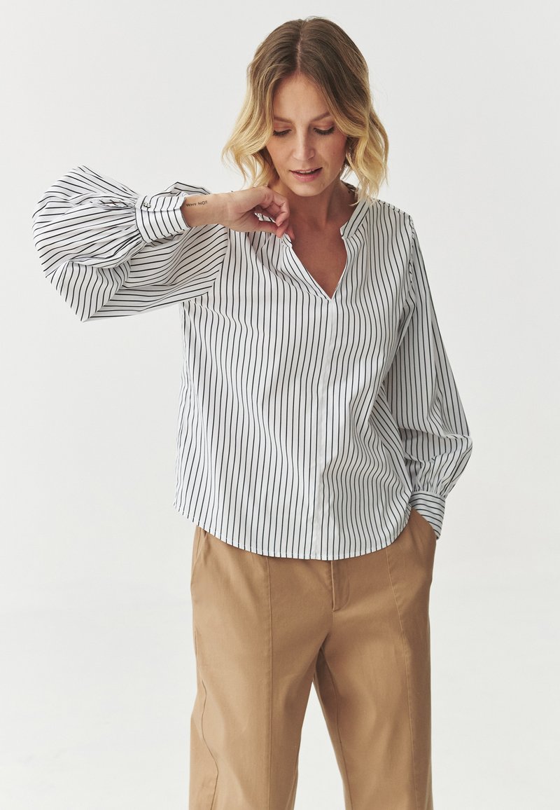 Camicia bianca con strisce verticali nere, scollatura a v e maniche lunghe a sbuffo. Abbinata a pantaloni beige a gamba larga. Tessuto morbido.