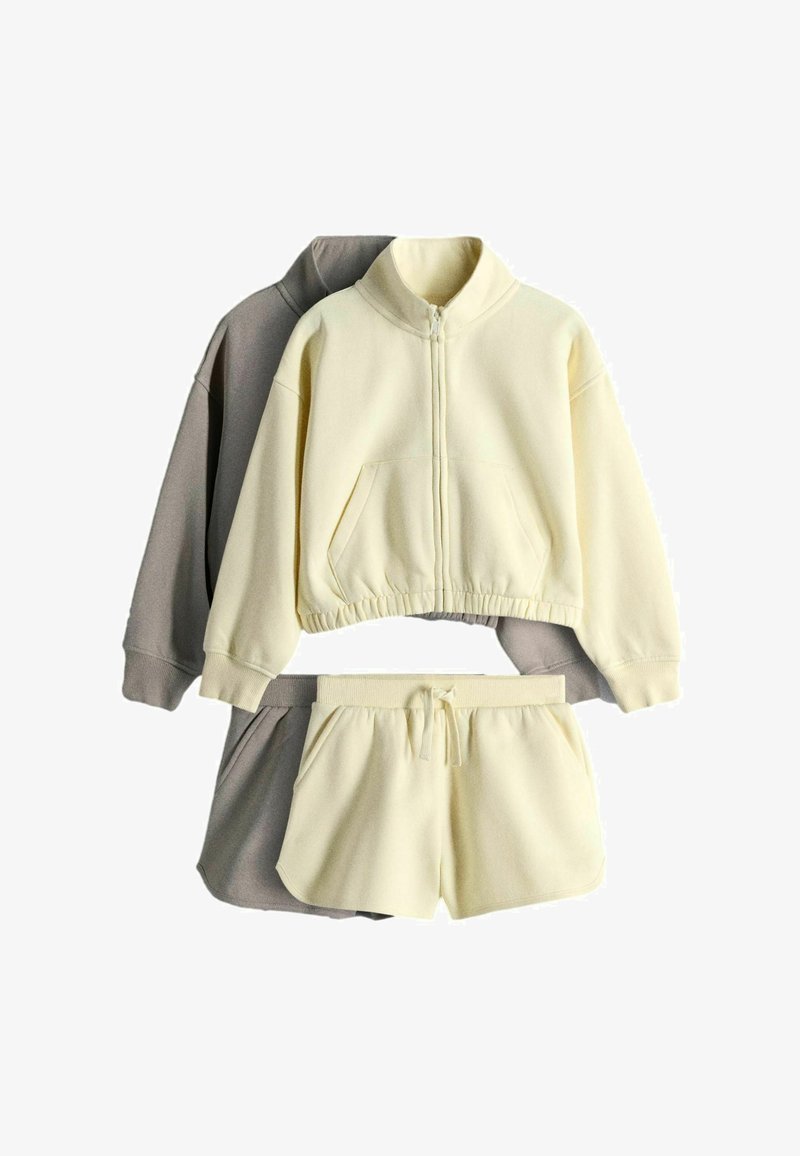 Twee sets fleecekleding met korte broeken met trekkoord en jasjes met rits, één in crème en de andere in taupe.