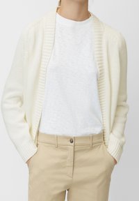 Persona che indossa un cardigan bianco a maglia, una t-shirt bianca a girocollo e pantaloni beige con le mani in tasca su uno sfondo neutro.