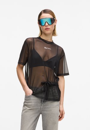 Kobieta w dużych lustrzanych okularach przeciwsłonecznych, przezroczystym czarnym siateczkowym topie na czarnym biustonoszu oraz jasnoszarych dżinsach, z jedną ręką w kieszeni.