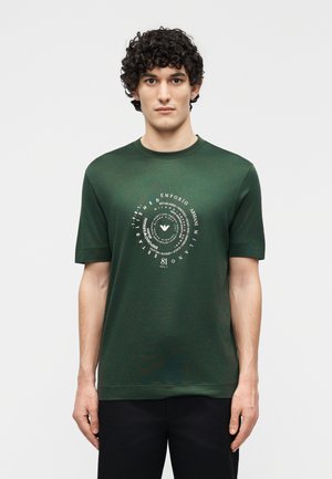 Άνδρας με σγουρά μαλλιά που φοράει μια κοντομάνικη μπλούζα σε σκούρο πράσινο χρώμα με κυκλικό λευκό λογότυπο Emporio Armani στο στήθος.